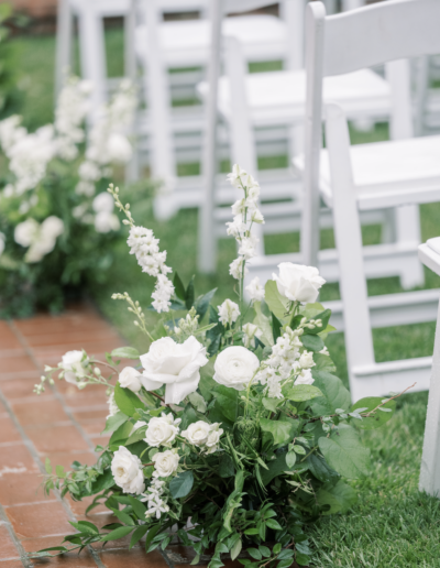 White flowers and greenery arranged beside white folding chairs on a brick path in an outdoor setting.